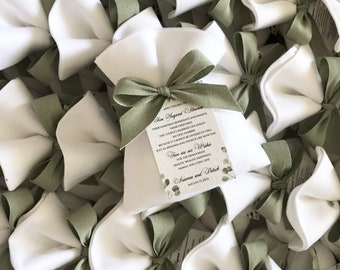 May include: A pile of white fabric pouches tied with green ribbon. Each pouch contains a small card with the text "Fire Sagared Moments" and a wedding date. The card also includes the text "Their happiness represents a chance, their tenderness, sweetness, simply married. The couple's wedding life ahead, an odd number, because it cannot be divided in two, just as Arianna and Patrick are now one. Give are our Wishes for the newlyweds, peace, love, health, wealth, happiness, family and long life. Arianna and Patrick, August 13, 2022."