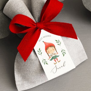 May include: A small gray burlap gift bag with a red ribbon tied around it. A white gift tag with the text "Best Wishes" and a watercolor illustration of a girl elf holding a bird is attached to the bag. The tag is signed "Sarah".