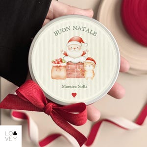 Peut inclure: Un cadeau de Noël rond avec un ruban rouge. Le cadeau a un bord argenté et présente une illustration du Père Noël avec un chat, un sac de cadeaux et le texte italien "Buon Natale". Les mots "Maestra Sofia" et un cœur rouge sont également présents.
