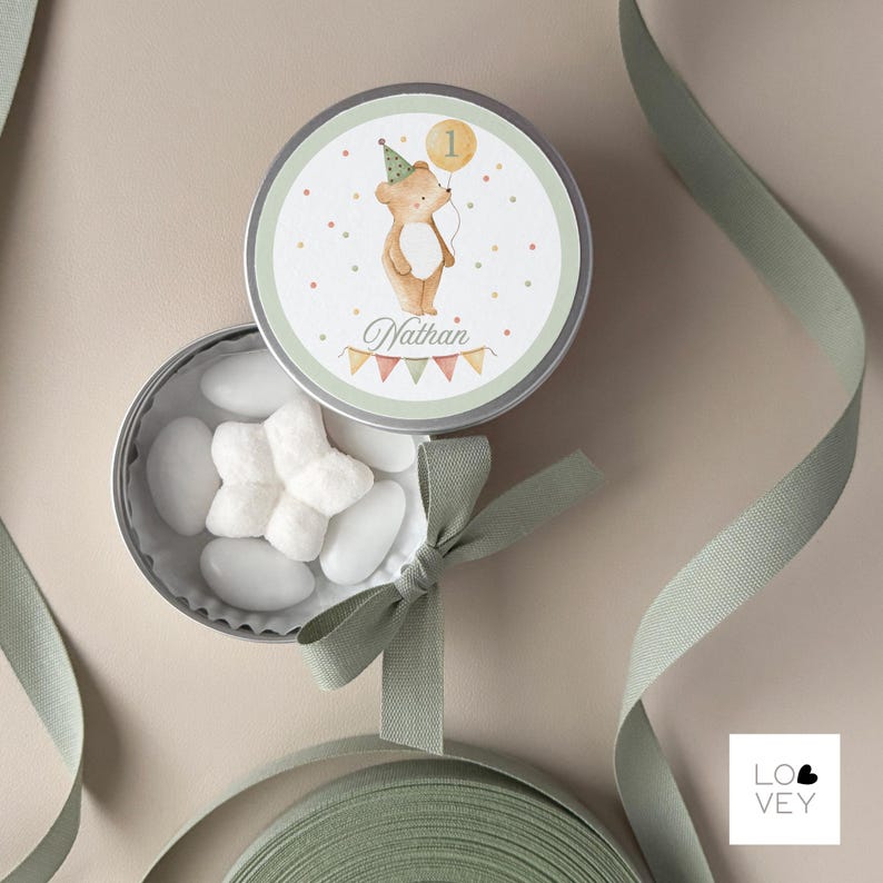 May include: A round, silver tin with a watercolor illustration of a bear holding a balloon with the number 1. The tin is open, revealing white candies. The tin lid has the name "Nathan" printed on it. A green ribbon and bow are also visible.