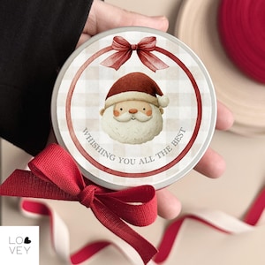 Peut inclure: Boîte de Noël ronde et blanche avec une illustration du Père Noël. La boîte présente un nœud rouge et le texte "Wishing you all the best" dans un design circulaire. Un ruban rouge est au premier plan.