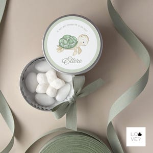 May include: A round, silver tin with a light green and white label featuring a cartoon turtle illustration. The tin is open, revealing white candies. A green ribbon and bow are also present.