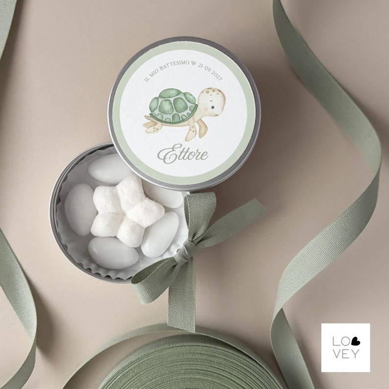 May include: A round, silver tin with a light green and white label featuring a cartoon turtle illustration. The tin is open, revealing white candies. A green ribbon and bow are also present.