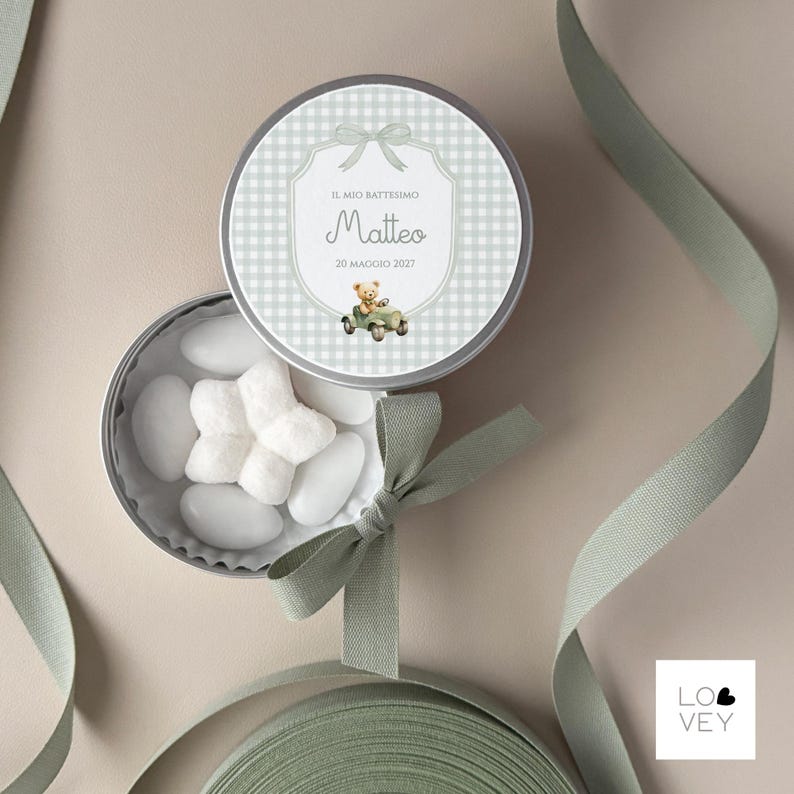 May include: A round, silver tin with a light green gingham pattern lid. The lid features the text "Il Mio Battesimo Matteo 20 Maggio 2027" and a teddy bear driving a car. The tin is open, revealing white candies and a green bow.