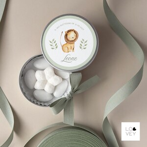 May include: A round, silver tin with a watercolor illustration of a lion cub, text that reads "Il mio battesimo 21 09 2022" and the name "Leone". The tin is open, revealing white candies. A green ribbon and bow are also present.