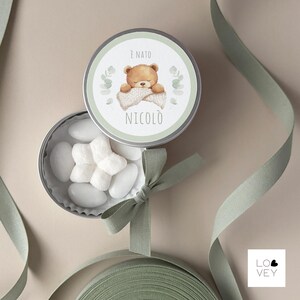 May include: A round, silver tin with a green and white label featuring a sleeping teddy bear illustration. The tin is open, revealing white candies and a green bow. The text "&Egrave; NATO NICOL&Ograve;" is printed on the label.