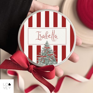 Op de afbeelding: Een ronde, zilveren blikken doos met een rood-witte gestreepte rand. Het midden heeft een witte rechthoek met de naam "Isabella" in cursief boven een grijze kerstboom met rode strikken. Een rode strik en lint staan op de voorgrond.