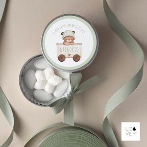 May include: A round, silver tin with a lid featuring a watercolor illustration of a teddy bear in a train car. The tin is open, revealing white candies. A green ribbon is tied around the tin. Text on the lid reads "IL MIO BATTESIMO 21.09.2021" and "GREGORIO".