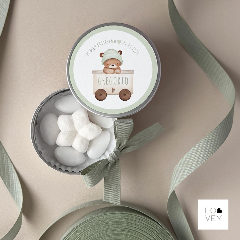May include: A round, silver tin with a lid featuring a watercolor illustration of a teddy bear in a train car. The tin is open, revealing white candies. A green ribbon is tied around the tin. Text on the lid reads "IL MIO BATTESIMO 21.09.2021" and "GREGORIO".