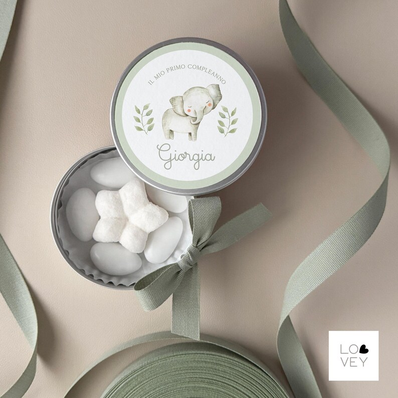 May include: A round, silver tin with a light green lid featuring a watercolor elephant illustration and the name "Giorgia". The tin is open, revealing white candies. A light green ribbon and bow are also present.