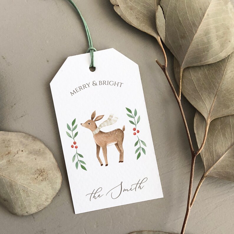 May include: White gift tag with a green string. The tag features a watercolor illustration of a deer wearing a scarf, surrounded by green leaves and red berries. The text "Merry & Bright" is printed at the top of the tag, and "The Smith" is printed at the bottom.