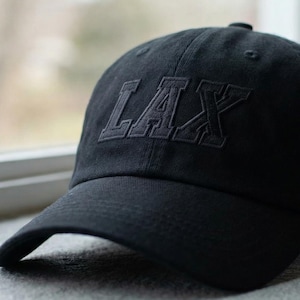 May include: A black baseball cap with the word "LAX" embroidered on the front. The cap is made of a soft material and has a curved brim. The cap is sitting on a gray surface.
