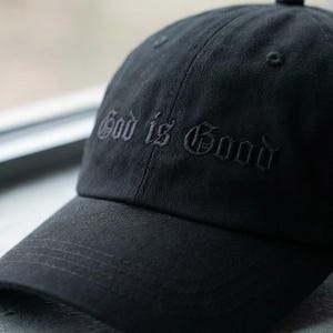 Könnte beinhalten: Schwarze Baseballkappe mit dem gestickten Schriftzug "God is Good" in gotischer Schrift. Die Kappe hat einen gebogenen Schirm und einen Knopf oben. Das Markenlogo "The Most Stitch" ist sichtbar.