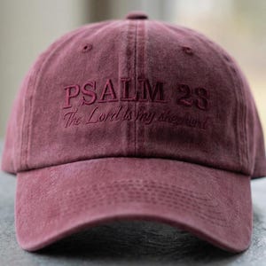 May include: A maroon baseball cap with the embroidered text "PSALM 23 The Lord is my shepherd." The cap has a curved brim and a fabric strap closure. The logo "THE MOST STITCH" is on the right side.