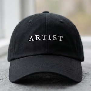 May include: A black baseball cap featuring the word "ARTIST" embroidered in white. The cap has a curved peak and a classic six-panel design. The hat is made of a soft, durable fabric, suitable for everyday wear.