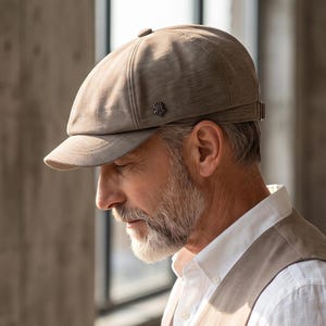 May include: A light brown baseball cap with a short, curved brim and a small metallic logo detail on the side. The cap features a structured crown with visible stitching and an adjustable strap at the back. The text THE MOST STITCH is visible.