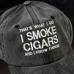 May include: A dark gray, distressed baseball cap with the embroidered text "THAT'S WHAT I DO I SMOKE CIGARS AND I KNOW THINGS." The hat is sitting on a wooden surface.