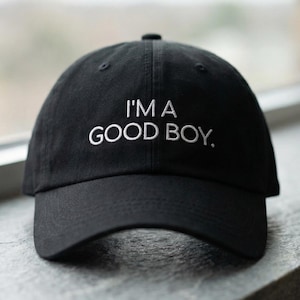Peut inclure: Casquette de baseball noire avec le texte "I'M A GOOD BOY." en blanc. La casquette a une visière incurvée et une couronne arrondie. Le chapeau est posé sur une surface grise.