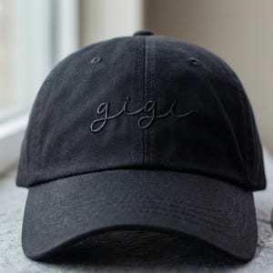 May include: A black baseball cap with the name "gigi" embroidered in a matching dark thread. The cap is on a window sill. The logo "THE MOST STITCH" is visible on the right side of the image.