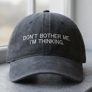 May include: A charcoal grey baseball cap with the white text "DON'T BOTHER ME. I'M THINKING." The cap is on a windowsill. It has a curved peak and a button at the crown. The cap is a casual accessory.