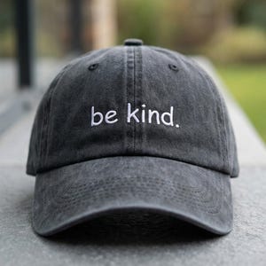 May include: A dark gray baseball cap with the embroidered phrase "be kind." The hat has a curved brim and a classic six-panel design. The text is in a clean, white font. The cap is made of a soft, textured material.