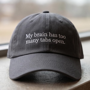 May include: A gray baseball cap with the embroidered text, "My brain has too many tabs open." The hat has a curved brim and a fabric strap closure. The logo "THE MOST STITCH" is visible on the right side.