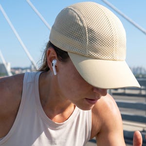 Puede incluir: Gorra de running beige de malla con visera curva. La persona lleva una camiseta sin mangas clara y auriculares inalámbricos blancos. El fondo muestra un puente y un cielo azul claro.