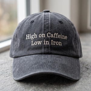 May include: A charcoal grey baseball cap with a distressed finish featuring the embroidered white text "High on Caffeine Low in Iron" centred on the front panel. The cap has a curved brim and a structured crown with visible stitching details.