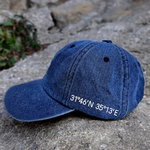 Può includere: Un cappello da baseball in denim blu con cuciture bianche. Il cappello ha le coordinate "31°46'N 35°13'E" ricamate sul retro.