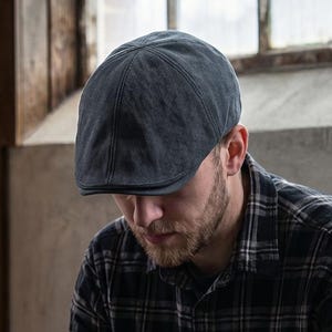 May include: A dark gray flat cap, worn by a person, paired with a black and gray plaid shirt. The image is set against a backdrop of wooden panels and a window, creating a rustic aesthetic. The text "THE MOST STITCH" is visible in the upper left corner.