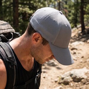 Op de afbeelding: Een grijze baseballcap met een mesh-ontwerp wordt gedragen door een persoon die op een pad wandelt. De cap heeft een gebogen klep en een gestructureerde kroon. Een rugzak is zichtbaar op de rug van de persoon. De tekst "THE MOST STITCH" is zichtbaar.