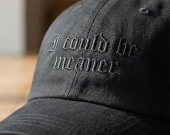 Può includere: Un cappellino da baseball nero con il testo ricamato "I could be meaner" in un carattere gotico sul pannello frontale. Il cappellino presenta una visiera curva, una corona strutturata con occhielli e un logo circolare "THEMOST STITCH" a sinistra.