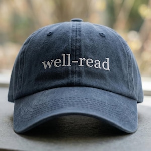 May include: A navy blue baseball cap with the embroidered text "well-read" in white. The hat is made of a soft material and has a curved peak. The Most Stitch logo is visible in the upper right corner.