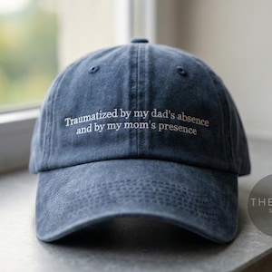 May include: A navy blue baseball cap with the embroidered text "Traumatized by my dad's absence and by my mom's presence." The cap is sitting on a gray surface near a window.