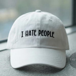 May include: White baseball cap with the embroidered phrase "I HATE PEOPLE" in black. The cap has a curved brim and a button on top. The text is centered on the front of the cap.