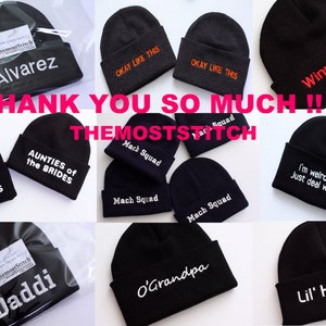 May include: A collection of black knit beanies with various embroidered designs, including "Alvarez", "Aunties of the Brides", "Mach Squad", "Daddi", "O'Grandpa", "I'm weird, ok? Just deal with it", "Winn/Dixie", "Lil' Homie", and "Okay Like This".