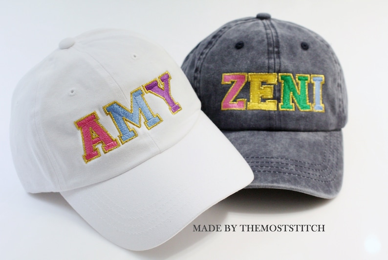 Op de afbeelding: Twee gepersonaliseerde baseballpetten, een witte met de naam "AMY" geborduurd in kleurrijke letters en een donkergrijze met de naam "ZENI" geborduurd in kleurrijke letters. De petten zijn gemaakt door TheMostStitch.