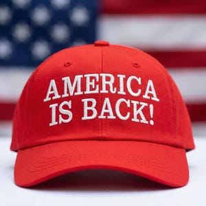 May include: A red baseball cap with the text "AMERICA IS BACK!" in white, set against a blurred American flag background. The cap is the primary focus, showcasing its bold message.
