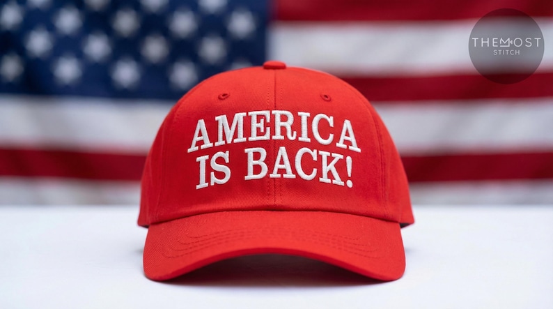 May include: A red baseball cap with the text "AMERICA IS BACK!" in white, set against a blurred American flag background. The cap is the primary focus, showcasing its bold message.