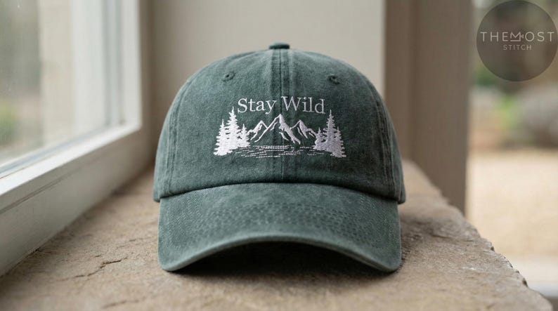 Puede incluir: Gorra de b&eacute;isbol verde oscuro con la frase bordada "Stay Wild" sobre un dise&ntilde;o de monta&ntilde;a y bosque blanco. La gorra est&aacute; hecha de un material suave y tiene una visera curva. La gorra est&aacute; sobre una superficie de piedra.