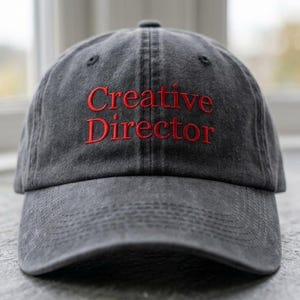 May include: A charcoal gray baseball cap with a washed, distressed finish. The front features the text Creative Director embroidered in red thread. The cap has a curved brim, a six-panel construction with eyelets, and a structured front crown.