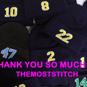 May include: A pile of navy blue knit beanies with various numbers embroidered on them in yellow and light blue. The numbers include 10, 8, 99, 47, 22, 14, 2, 1, and 144. The text "THANK YOU SO MUCH!! :) THEMOSTSTITCH" is visible at the bottom of the image.