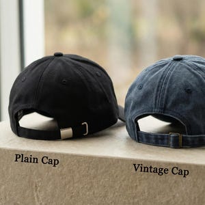 Puede incluir: Dos gorras de b&eacute;isbol, una negra y otra azul descolorida, se muestran juntas. La gorra negra tiene una hebilla plateada, y la azul una hebilla bronce. El texto "Plain Cap" y "Vintage Cap" est&aacute; debajo.