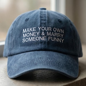 Könnte beinhalten: Blaue Denim-Baseballkappe mit dem gestickten Text "MAKE YOUR OWN MONEY & MARRY SOMEONE FUNNY". Die Kappe hat einen gebogenen Schirm und einen Stoffriemenverschluss.