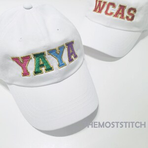 Op de afbeelding: Witte baseballpet met een kleurrijk geborduurd woord "YAYA" op de voorkant. De pet heeft een gebogen klep en een witte verstelbare band.
