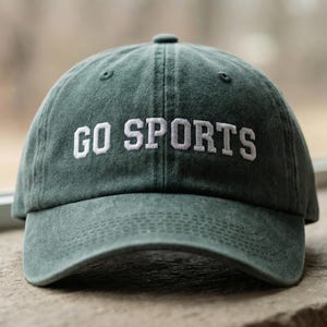 May include: A dark green baseball cap with a washed, vintage-style finish. The front of the cap features the text GO SPORTS embroidered in white capital letters. The hat has a curved brim and a structured crown with visible stitching details.