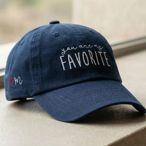 May include: Navy blue baseball cap with the embroidered text "you are my FAVORITE" in white. A small red heart and the letter "M" are embroidered on the side. The cap is sitting on a stone surface.