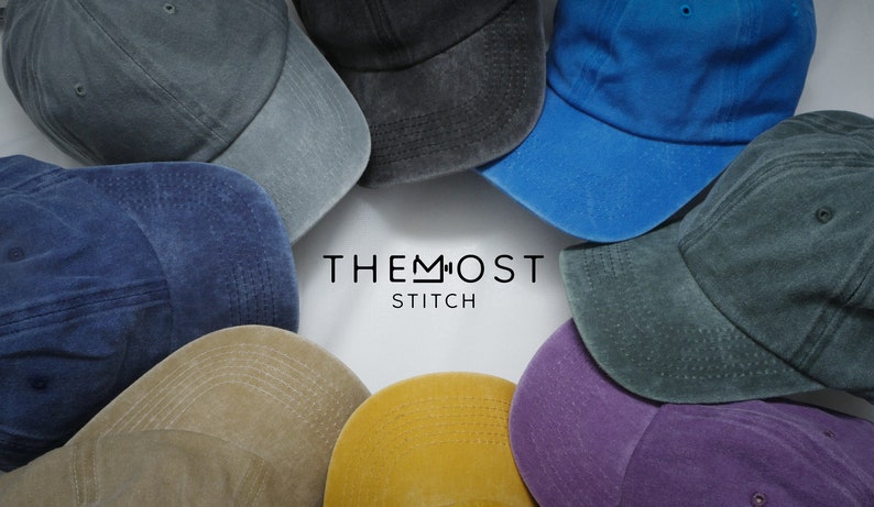 Op de afbeelding: Een verzameling van negen baseballpetten in verschillende kleuren, waaronder blauw, grijs, groen, paars, geel en bruin. De petten zijn in een cirkel rond de tekst "THE MOST STITCH" gerangschikt.