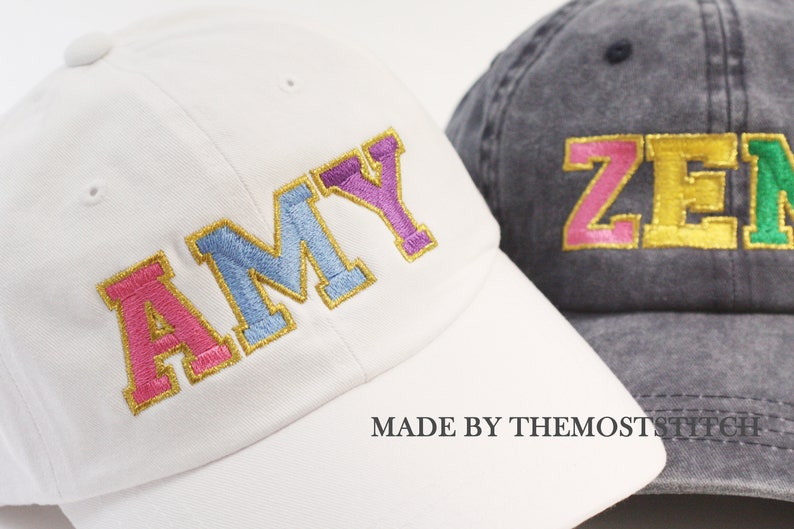 Op de afbeelding: Twee baseballpetten met geborduurde letters. De witte pet heeft de naam "AMY" in roze, blauwe en paarse letters met een gouden omlijsting. De grijze pet heeft de naam "ZEN" in roze, gele en groene letters met een gouden omlijsting. Beide petten hebben de tekst "MADE BY THEMOSTSTITCH" onderaan.