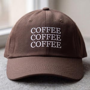 May include: A brown baseball cap featuring the word COFFEE embroidered three times in white capital letters stacked vertically on the front panel. The cap has a structured crown, a curved brim, and a button at the top centre of the dome.
